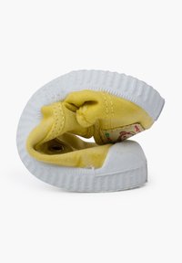 Zapatilla de tela amarilla con suela de goma blanca, que presenta un diseño plegado y un pequeño parche decorativo con un personaje de oso en el lateral.