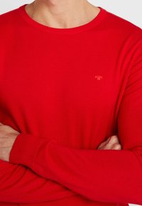 Pull-over rouge à manches longues en matière côtelée. Il présente un col rond et un petit logo dans une teinte plus claire sur la poitrine.