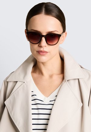 Donna con occhiali da sole sfumati scuri, trench color beige e camicia bianca a righe orizzontali nere, guarda avanti con espressione neutra.