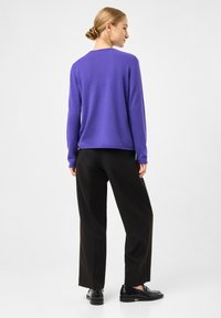 Kvinde med blond hår i en knold iført en lilla langærmet top, sorte bukser og sorte loafers, står med ryggen til på en ensfarvet baggrund.