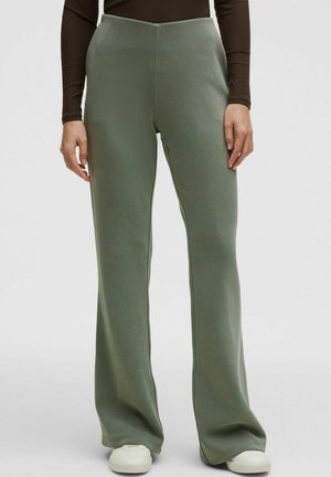 Žena nosící olivově zelené široké kalhoty a bílé boty stojí před jednoduchým světlým pozadím.