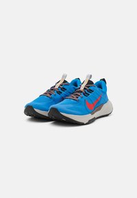 Buty biegowe Nike w intensywnym niebieskim kolorze, z czerwonym swooshem, szarym podeszewką, czarnymi akcentami oraz teksturowaną siateczką na cholewce i brązowymi sznurówkami.