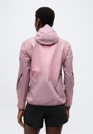 Personne portant une veste à capuche violet clair et un short noir, debout dos à la caméra sur un fond uni.