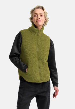 Frau trägt olivgrüne Fleeceweste mit schwarzem Langarmshirt und schwarzer Hose, steht mit einer Hand in der Tasche vor weißem Hintergrund.