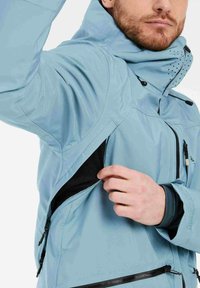 Lichtblauwe waterdichte jas met ritszakken, verstelbare capuchon en textuurstof. Heeft een zwarte gaasvoering en ergonomisch ontwerp.