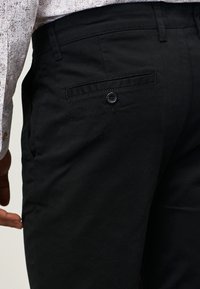 Pantaloni in cotone neri con fronte piatto, tasche laterali e una tasca posteriore singola con chiusura a bottone. Tessuto liscio e vestibilità su misura.