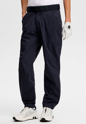 Marineblaue Golfhose aus leichtem Stretchstoff. Mit Gürtel, Seitentaschen und schmaler Passform. Kombiniert mit weißen Sportschuhen.