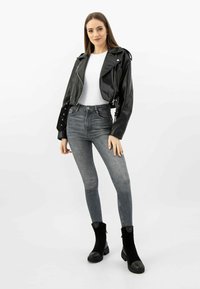 Žena v černé zkrácené kožené bundě, bílé košili, šedých skinny džínách a černých kotníkových botách, stojící na bílém pozadí.