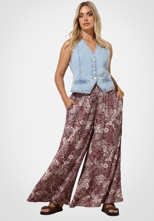 Vrouw die een mouwloze lichtblauwe spijkervest draagt, bordeauxrode palazzo-broek met bloemenprint en zwarte sandalen, staand met handen in de zakken.