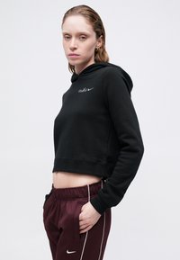 Melna īsā kapucēta jaka ar savilcēju un izšūtu Nike logo. Uzvilkta ar vīna krāsas treniņa biksēm ar baltām sānu svītrām.