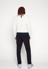 Vit kabelstickad tröja, mörka byxor och silvriga sportskor. Tröjan har en kort, boxig passform med en blå detalj vid nederkanten.