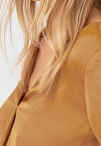 Blusa in satin giallo senape con scollatura a V e texture morbida. Presenta una vestibilità comoda con maniche fluide e una finitura liscia.