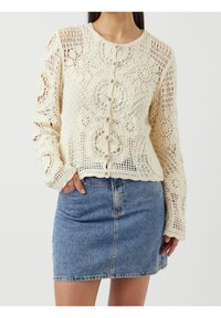 Szydełkowy kardigan w kremowym kolorze z kwiatowymi wzorami i guzikami z przodu, połączony z dżinsową mini spódnicą. Posiada długie rękawy i otwarty splot.
