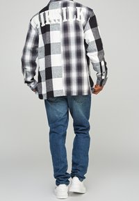 Camisa de franela a cuadros en blanco y negro con grandes letras "SikSilk" en la parte de atrás, combinada con jeans azules y zapatos blancos.