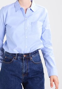 Camicia azzurra chiara con colletto e taschino, abbinata a jeans in denim blu scuro a vita alta con un classico design a cinque tasche.