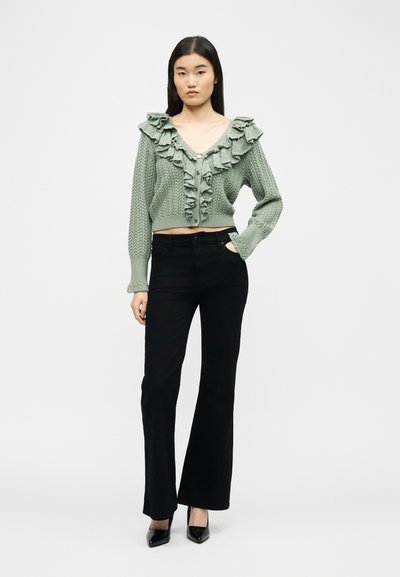 Jonge vrouw die een saliegroene gerimpelde gebreide cardigan draagt en zwarte wijde broek met zwarte puntige hakken, staande tegen een effen witte achtergrond.