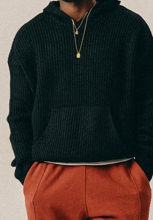 Personne portant un sweat à capuche en maille noire, un pantalon couleur rouille et deux colliers en or, debout les mains dans les poches devant un fond uni.