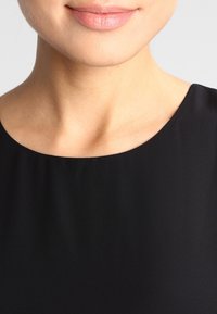 Blouse noire avec une texture lisse et mate, dotée d'un col rond, présentant des lignes simples et épurées sans ornements.