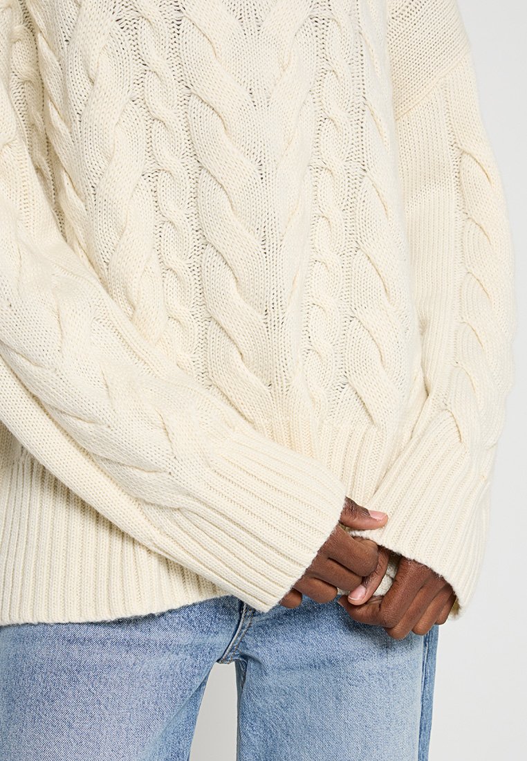 Cremfarbener Kabelstrickpullover mit strukturiertem Muster, gerippten Bündchen und Körper, getragen über hellblauen Jeans. Weicher, voluminöser Strickstoff.