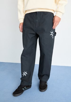 Pantalones negros de corte holgado, hechos de un tejido duradero y con dos bolsillos. Presentan un detalle de estrella bordada blanca en la pierna izquierda. Se llevan con zapatillas oscuras.