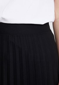 Svart plisserad kjol med en slät elastisk midjeband. Tyget har en mjuk textur, med vertikala veck och en neutral-färgad topp.