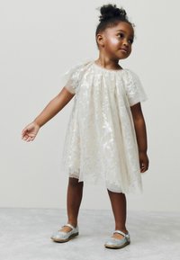 Robe courte sans manches avec une superposition transparente couverte de motifs d'étoiles en argent ; couleur de base crème ; associée à des chaussures à paillettes argentées.