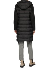 Zwarte puffer vest met een capuchon, voorzien van gestikte secties en een bovenkant van zachte wolmix. Gecombineerd met lichtgrijze broek en beige enkellaarsjes.