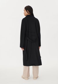 Lång svart kappa med bältad midja, krage och långa ärmar. Tyget verkar slätt med en strukturerad form. Ljusfärgade byxor och skor syns.