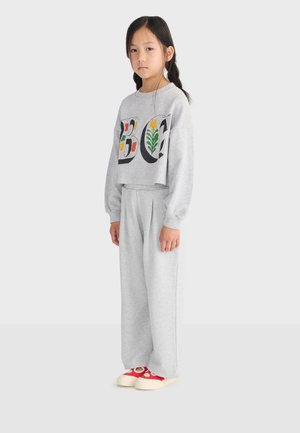 Jong meisje met zwart gevlochten haar, draagt lichtgrijze trainingsbroek en een bijpassend sweatshirt met bloemmotieven in de letters, staand in zijaanzicht.