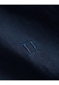 Tummansininen pellavakangas, jossa on hienovarainen tekstuuri ja kirjailtu "II" samansävyisellä sinisellä langalla, mikä lisää hienostuneen yksityiskohdan.