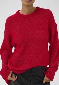 Punane kootud kampsun, millel on ümmargune kaelus, pikad varrukad laia mansetiga ja lai lõige. Tekstuur on pehme ja veidi läikiv.