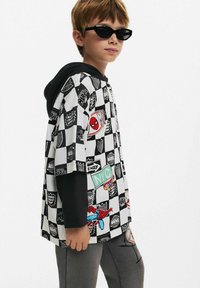 Kortärmad hoodie i svart och vit rutmönster med Marvel-grafik, röda detaljer och en svart huva. Grå byxor kompletterar outfiten.