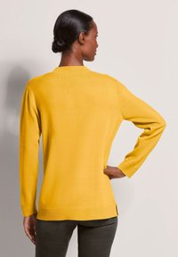 Gelber gestrickter Pullover mit langen Ärmeln, rundem Halsausschnitt, gerippten Bündchen und seitlichen Schlitzern am Saum. Glatte Textur, lockere Passform.