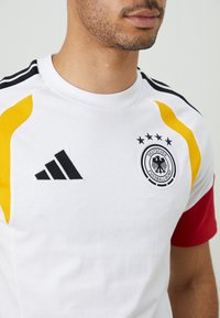 Camiseta atlética branca com detalhes em preto, amarelo e vermelho. Apresenta um decote redondo, textura em malha e um logótipo circular da federação de futebol com estrelas.