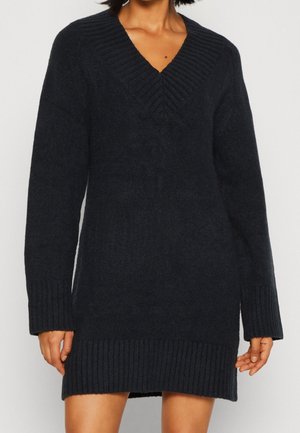 Frau, die ein langes, schwarzes, gestricktes Sweaterkleid mit tiefem V-Ausschnitt und gerippten Bündchen und Saum trägt.