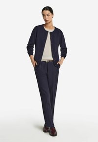 Granatowy sweter z dzianiny z okrągłym dekoltem i zapięciem na guziki, noszony na kremowej bluzce, połączony z granatowymi spodniami i błyszczącymi burgundowymi butami.