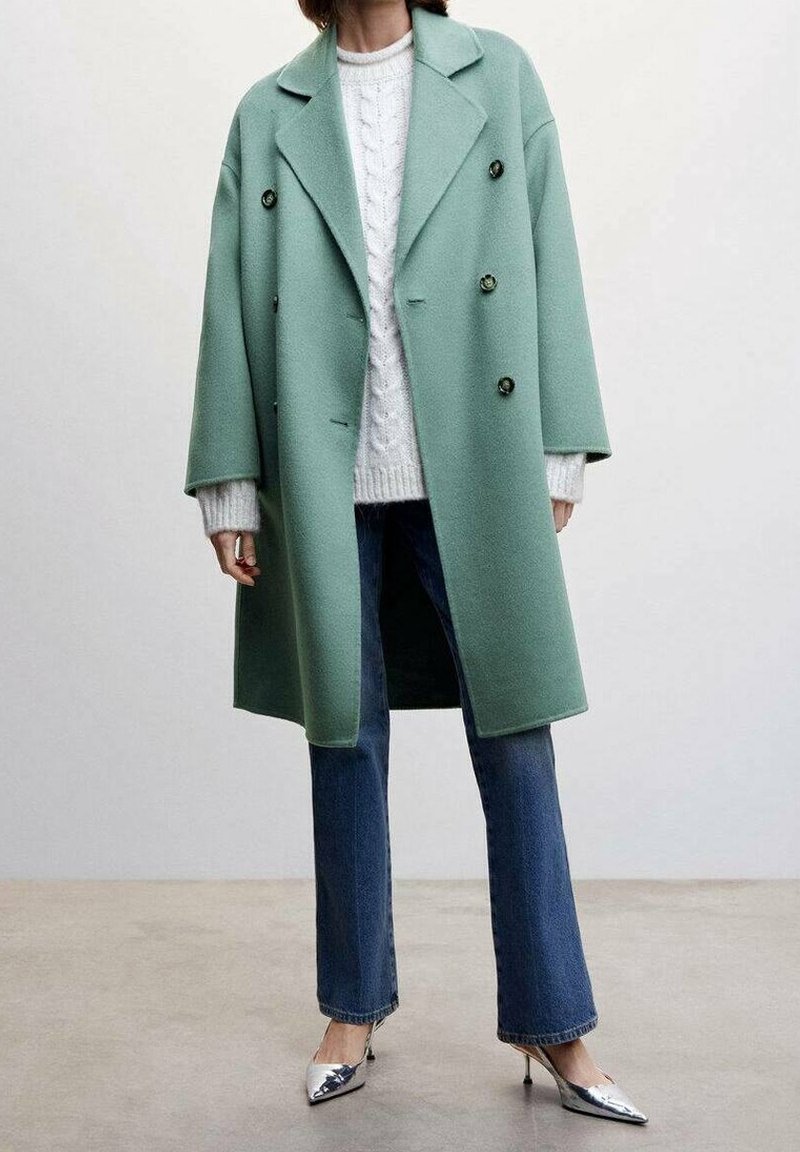Manteau en laine vert menthe avec un design croisé et de grands boutons, porté sur un pull en tricot à torsades blanc et un jean bleu, associé à des talons argentés.