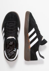 Zapatillas de ante negro con franjas de cuero blanco, puntera redonda, suela de goma marrón texturizada y cuello acolchado para mayor comodidad.