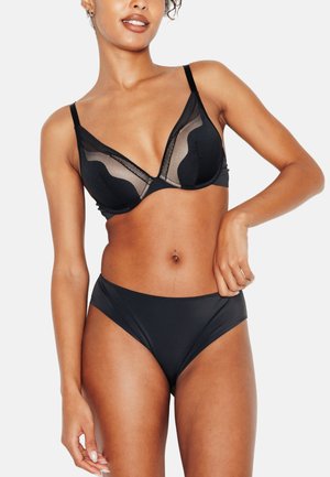 Femme portant un soutien-gorge noir transparent et opaque avec une culotte noire taille haute assortie, posant avec une main sur la hanche devant un fond blanc.