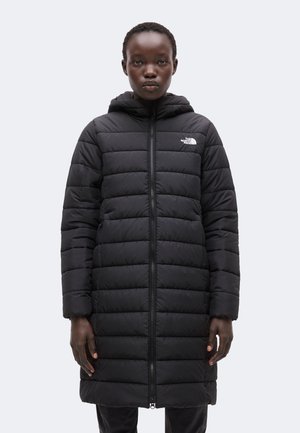 Person iført en lang sort quiltet hættejakke med lynlås foran og The North Face-logo på venstre bryst, stående mod en ensfarvet baggrund.