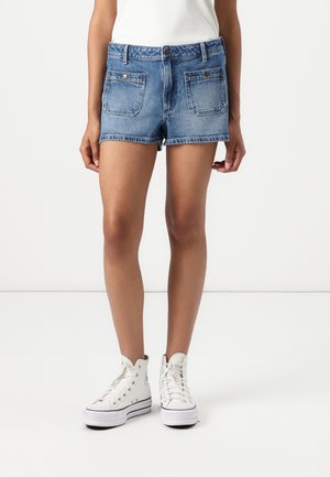 Personne portant un short en jean bleu avec des poches avant et des baskets blanches montantes, debout sur un sol blanc avec un fond uni.
