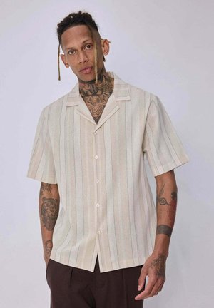Homme tatoué portant une chemise beige à manches courtes avec des rayures verticales et un pantalon foncé, debout devant un fond clair uni.