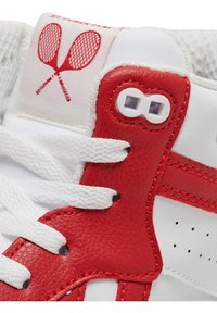 Hvide sneakers med røde accenter, der har et struktureret læderpanel, fremtrædende snøreringe og en grafik af krydsede ketsjere på tungen.