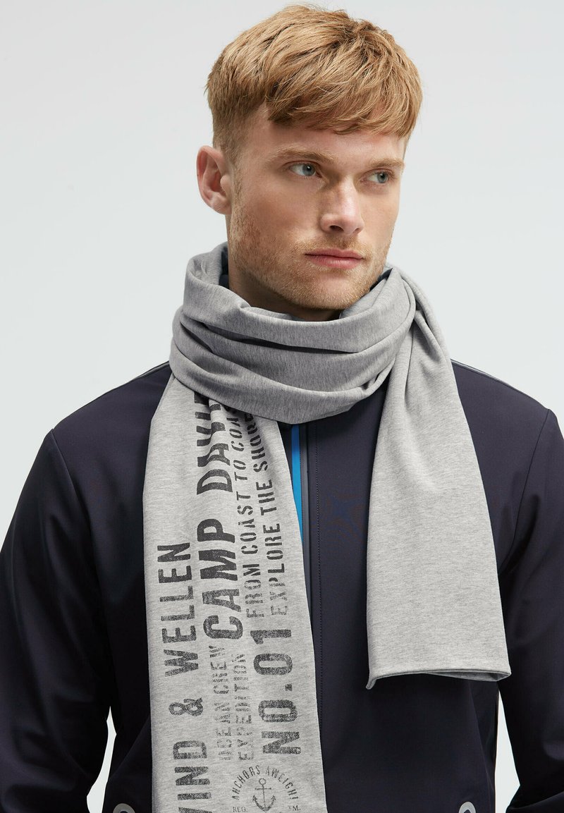 Young man with short red hair wearing a navy jacket and a light gray scarf with black printed text around his neck.