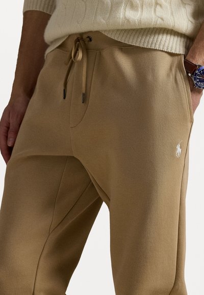 Beige sporta bikses ar auklu jostu, sānu kabatām un mazu baltu polo logotipu uz augšstilba, izgatavotas no mīksta, ērtā materiāla.
