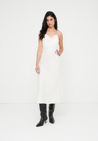 Λευκό μίντι φόρεμα χωρίς μανίκια με λαιμόκοψη καρδιά, διαθέτει ζώνη που δένει στη μέση και λείο, ρέον ύφασμα. Μαύρες μπότες με ψηλό τακούνι.