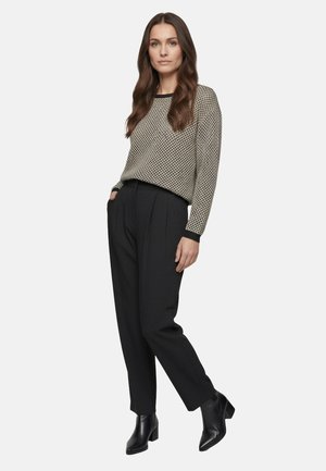 Frau mit langen braunen Haaren, die schwarze Knöchel-Stiefel, schwarze Hose mit hoher Taille und einen beige gemusterten langärmeligen Pullover trägt, steht mit einer Hand in der Tasche.