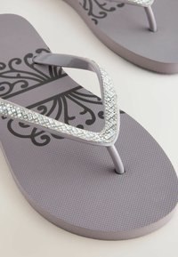 Infradito viola con superficie strutturata, caratterizzati da un prominente motivo decorativo nero e accenti scintillanti di strass sulle cinghie.