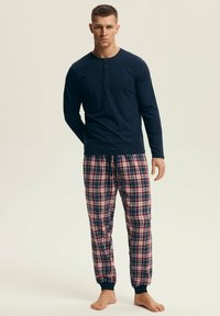 Mężczyzna w granatowej, długorękawowej koszulce henley i czerwono-biało-granatowych flanelowych spodniach do piżamy, stojący boso na beżowym tle.