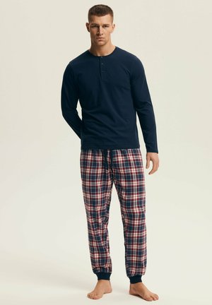 Mężczyzna w granatowej, długorękawowej koszulce henley i czerwono-biało-granatowych flanelowych spodniach do piżamy, stojący boso na beżowym tle.